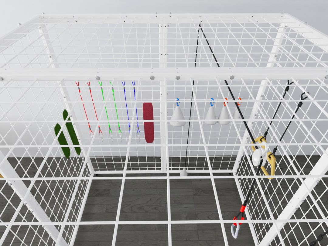 Overhead view of the Summit Sensory Gym Universal Exercise Unit, showcasing its white metal grid structure and interior accessories. The cage contains a variety of therapy tools, including colorful resistance bands, a padded red support, white cone-shaped accessories with hooks, and yellow handles attached to ropes. The grid design provides versatility for attachment points, making it suitable for different therapy exercises. The setup is displayed on a dark wood-style floor in a clean, professional environ