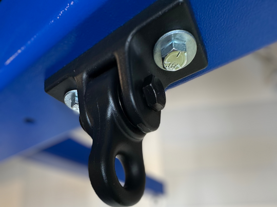 Close-up view of the Summit Soar Free Standing Swing Frame's heavy-duty connection point. The image highlights a black steel swivel hook securely mounted to a vibrant blue frame with robust bolts and washers. The durable design ensures safety and flexibility for swing attachment. In the background, part of the sensory club environment is visible, featuring a "Toddler Area" sign and a mural of rolling green hills.